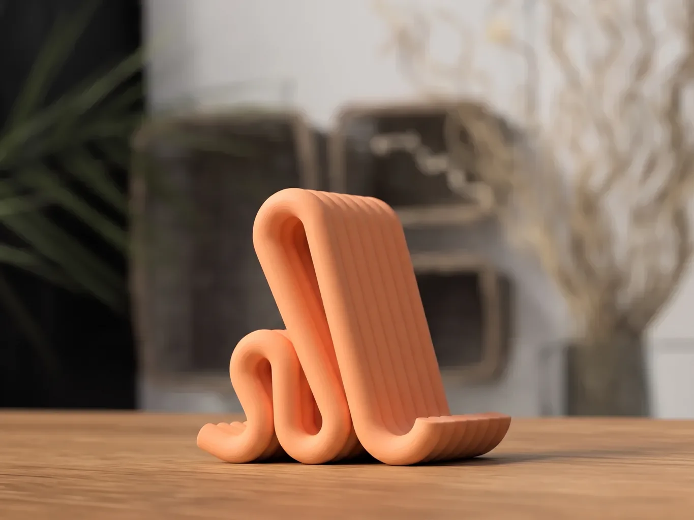 Support de téléphone imprimé en 3D, ruban nervuré replié en boucle, coloris orange mat, posé sur un bureau en chêne