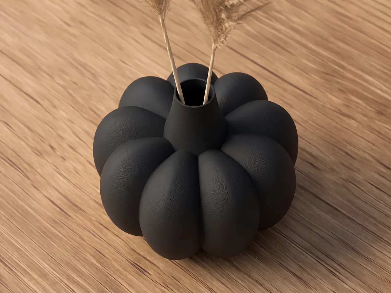 3D printed pumpkin vase in matte black, body with large rounded ribs and narrow neck, filled with two stems of dried grasses, on an oak table
